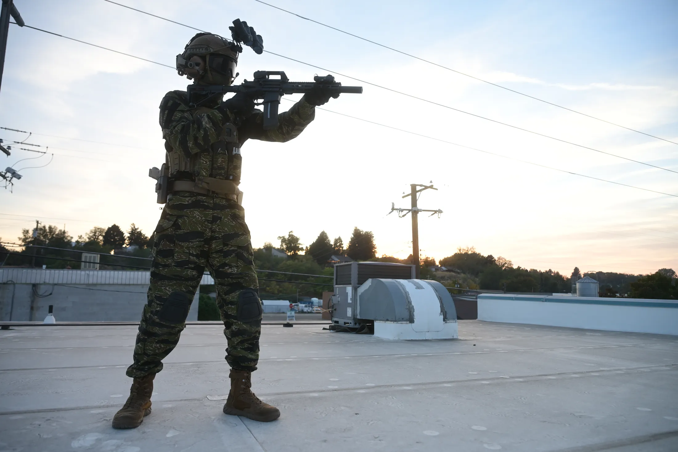 Wide shot of the commando aiming from a rooftop at golden hour, from Rusty Rehl's Combat Sport Supply ad