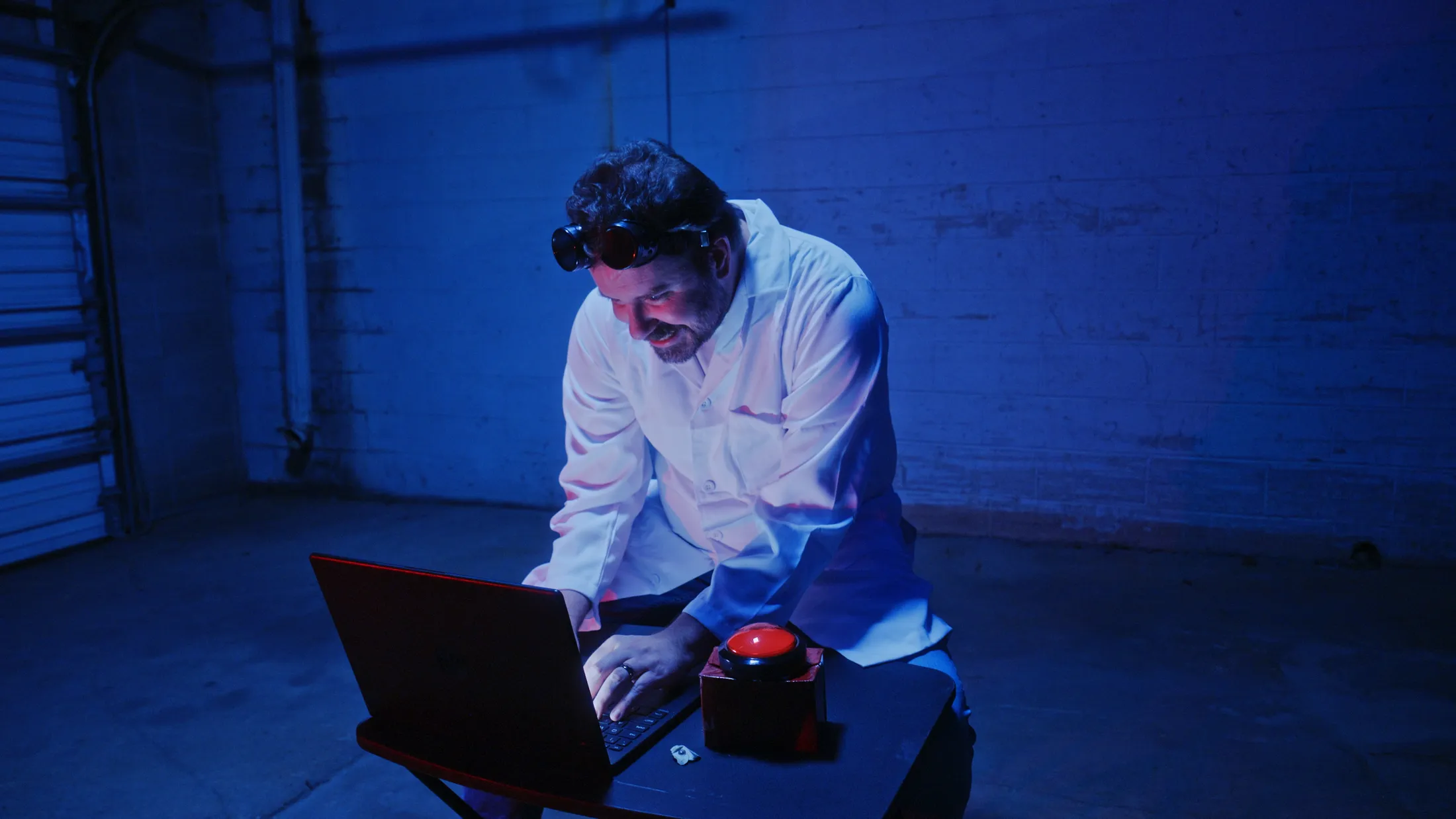 Mad scientist typing at a red laptop beside a big red button in a blue-lit room, still from Rusty Rehl's Combat Sport Supply ad