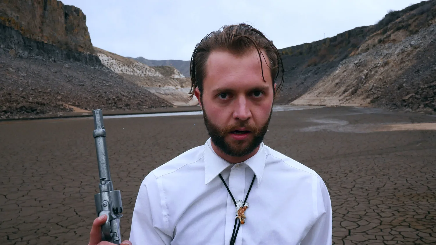 Still from a Rusty Rehl short film: a bearded man in a white shirt and bolo tie holds a revolver on a cracked, dry reservoir bed in Idaho