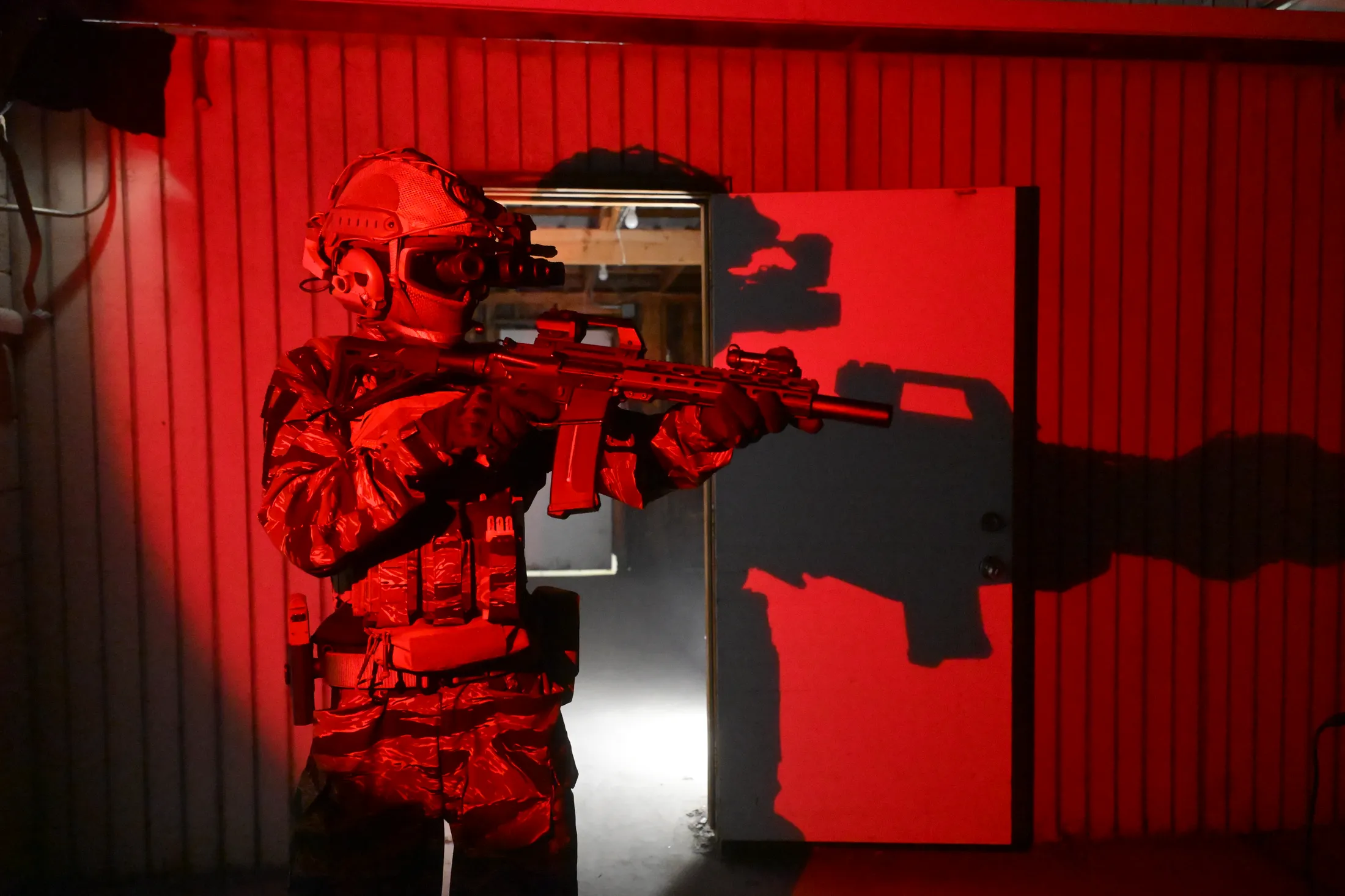 Commando in tiger-stripe camouflage and night-vision goggles holding a rifle, bathed in red light with his shadow on an open door, photo by Rusty Rehl for a Combat Sport Supply ad