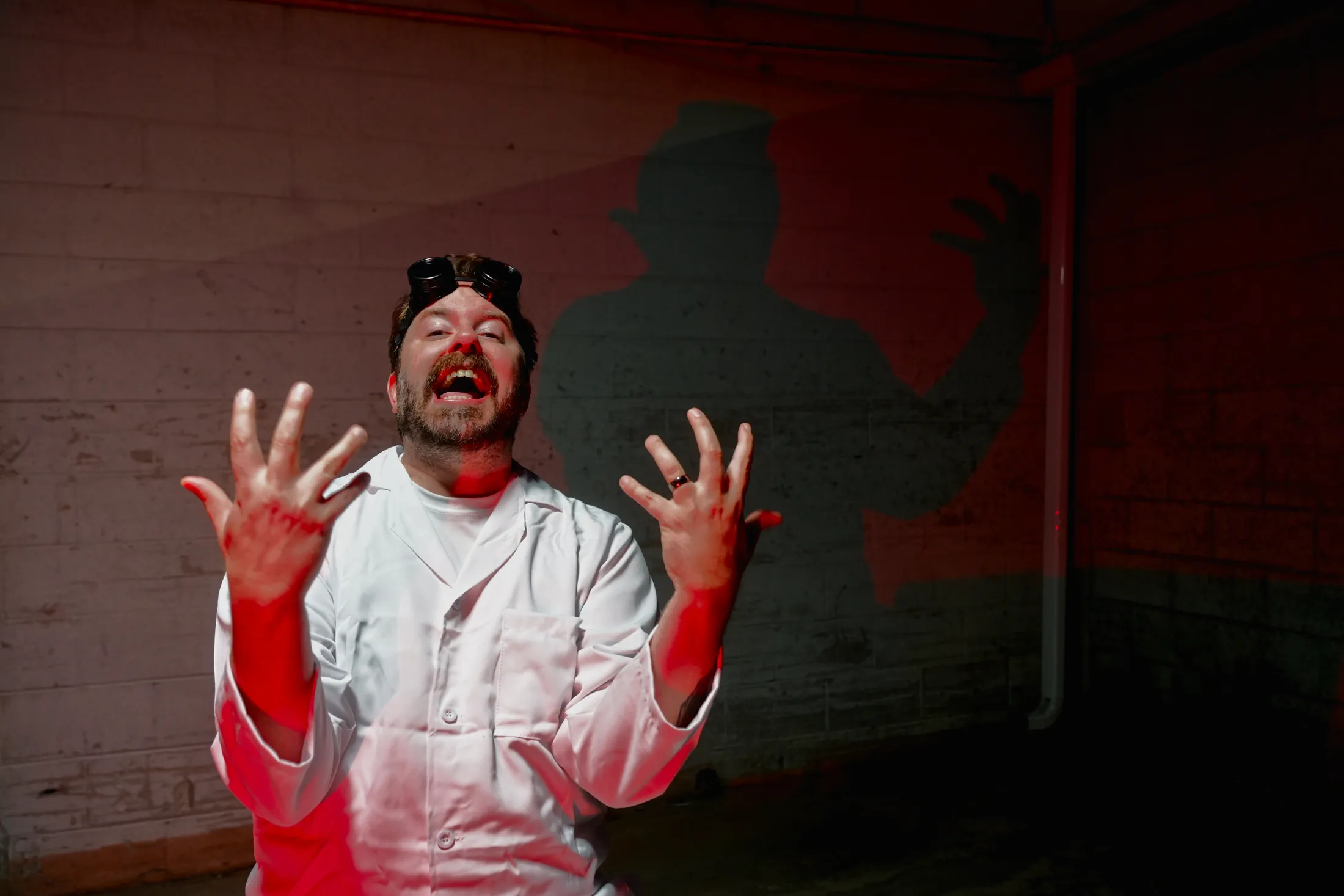Mad scientist in a lab coat with goggles raising his hands in red light, his shadow cast on a cinderblock wall, photo by Rusty Rehl for a Combat Sport Supply ad