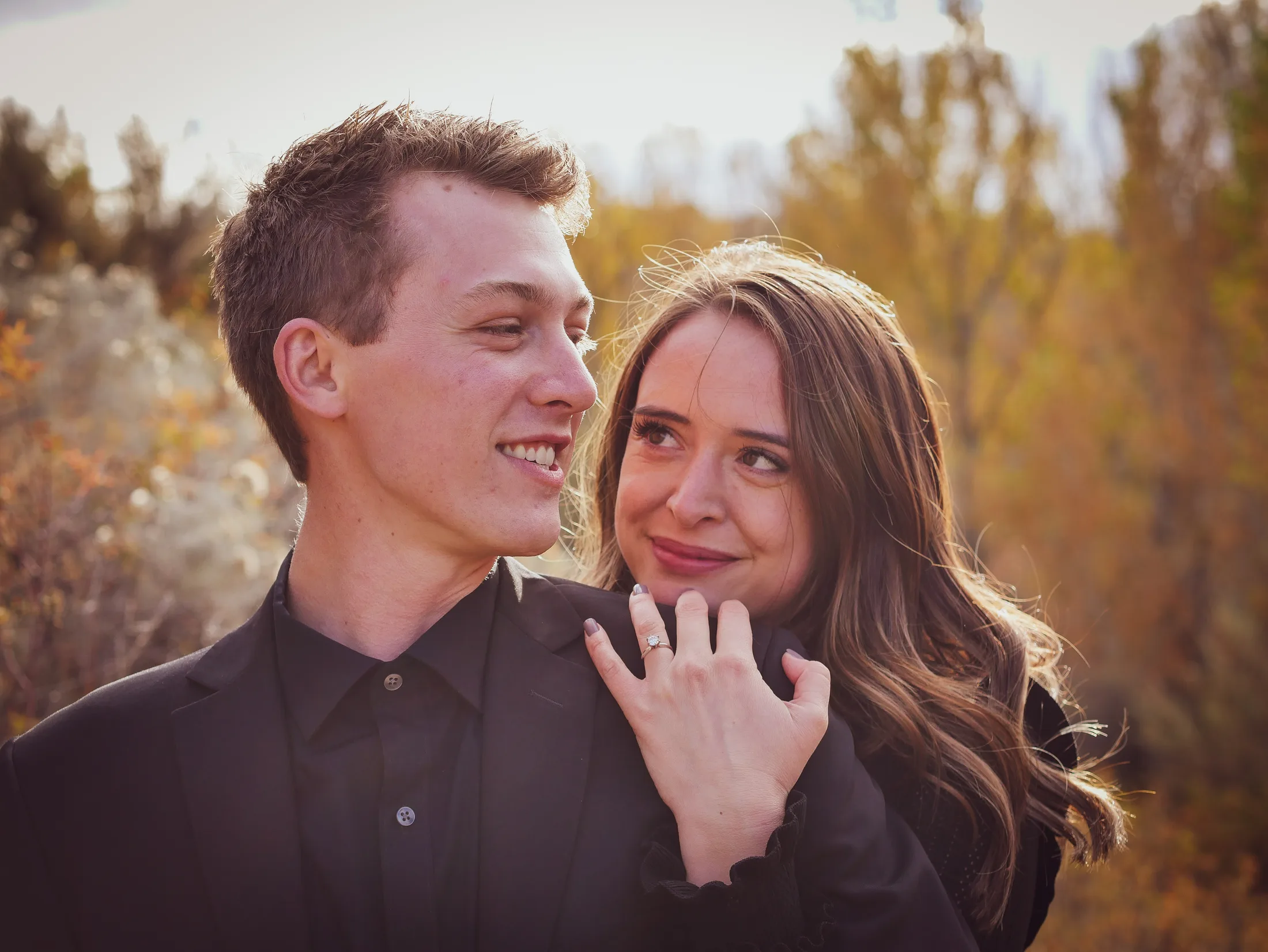 Engaged couple in fall color, she rests her hand with the engagement ring on his shoulder as they smile at each other, Boise, Idaho engagement photo by Rusty Rehl