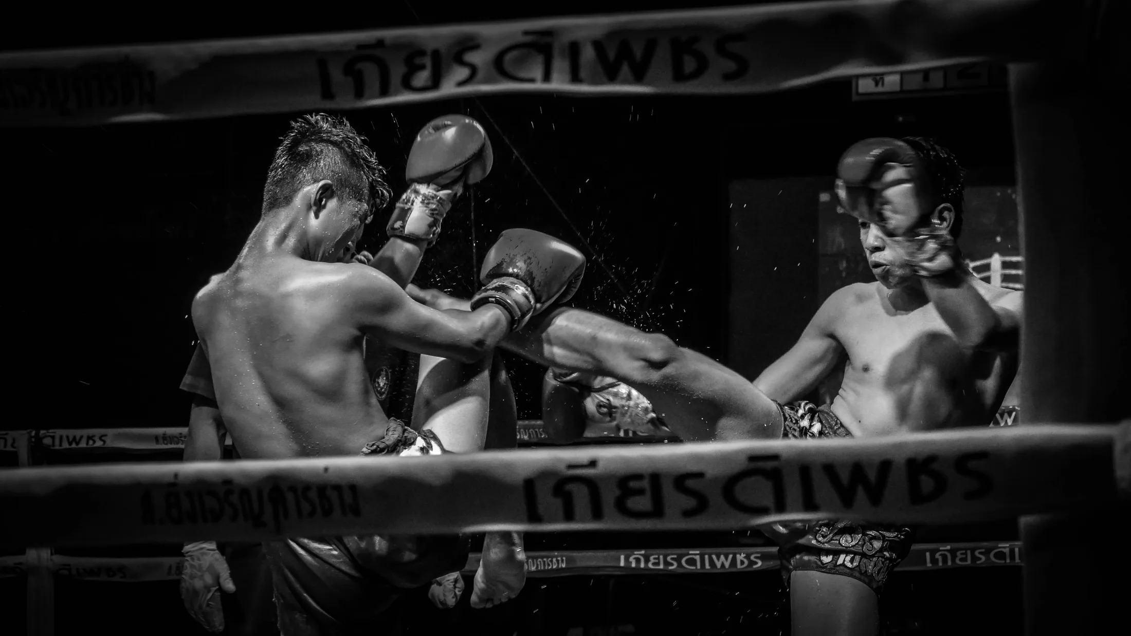 Muay Thai fighters mid-kick inside the ring ropes, black-and-white fight photo from Thailand by Rusty Rehl