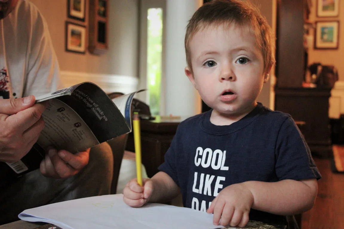 Toddler in a Cool Like Dad t-shirt holding a pencil and looking up from his drawing, candid family photo by Boise, Idaho photographer Rusty Rehl
